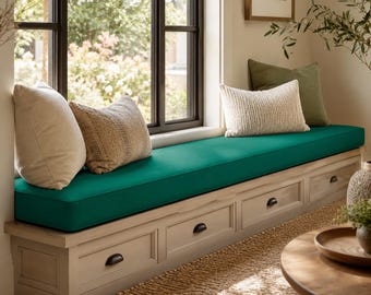 May include: A window seat bench featuring a long, solid teal cushion and three storage drawers with dark metal pulls. The bench is topped with two cream-colored pillows and one textured beige pillow, positioned against a large window frame.
