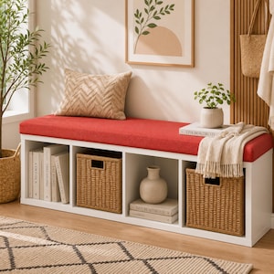May include: A white storage bench with a red cushioned seat featuring four cubby compartments. Two cubbies contain woven wicker baskets, one holds a white ceramic vase on a book, and one holds a row of books. A chevron-patterned pillow sits on top.