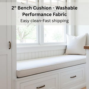 May include: A 2-inch bench cushion made of washable performance fabric. The cushion is rectangular, with a light beige border and a white center. It is placed on a white bench with drawers, near a window. The text on the image says "2" Bench Cushion - Washable Performance Fabric".