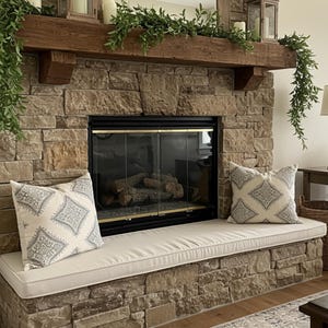 May include: A stone fireplace with a black framed glass door. Two decorative pillows with a gray and white pattern sit on a cream-colored bench. A wooden mantel is decorated with greenery and candles.