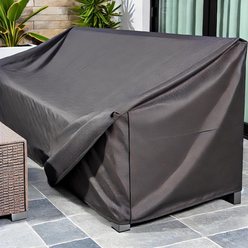 High-quality dark grey cover, tightly fitted over an outdoor sofa set. The water-resistant fabric offers protection from the elements, with a stone patio and potted plants completing the elegant, functional outdoor setting.