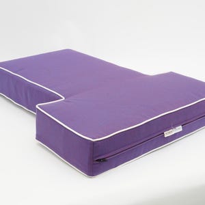 May include: A purple L-shaped cushion with white piping and a zipper on one side.