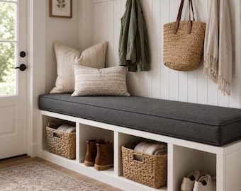May include: A white entryway bench with a dark gray cushion, featuring four storage cubbies containing two woven baskets, a pair of brown boots, and a pair of white sneakers. Above the bench are hooks holding a jacket, a straw tote, and a scarf.