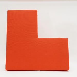 May include: An orange, L-shaped, upholstered furniture piece. The item is a vibrant shade of orange and appears to be made of a soft, textured fabric. The design is modern and minimalist, with clean lines and a simple shape.