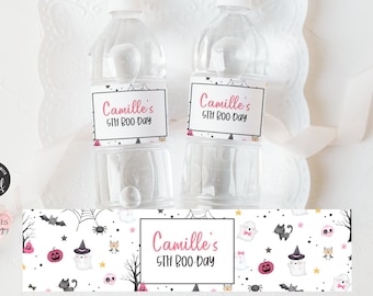 Może przedstawiać: Dwie przezroczyste plastikowe butelki z wodą z białymi zakrętkami i etykietami z napisem "Camille's 5th Boo Day". Poniżej butelek znajduje się dekoracyjny baner z halloweenowymi ilustracjami duchów, dyni i nietoperzy.
