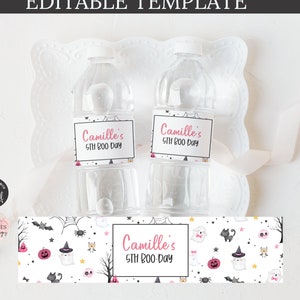 May include: Two clear plastic water bottles with white lids and labels saying "Camille's 5th Boo Day." A Halloween-themed banner with ghosts, pumpkins, and bats is below the bottles. The bottles are on a white surface.