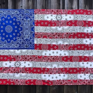 May include: A large American flag made from red, white, and blue bandanas. The flag is hanging on a wooden fence.