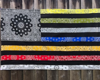 May include: A black and white bandana flag with stripes of yellow, green, gray, blue, red, and white. The flag is made of fabric and has a patchwork design.