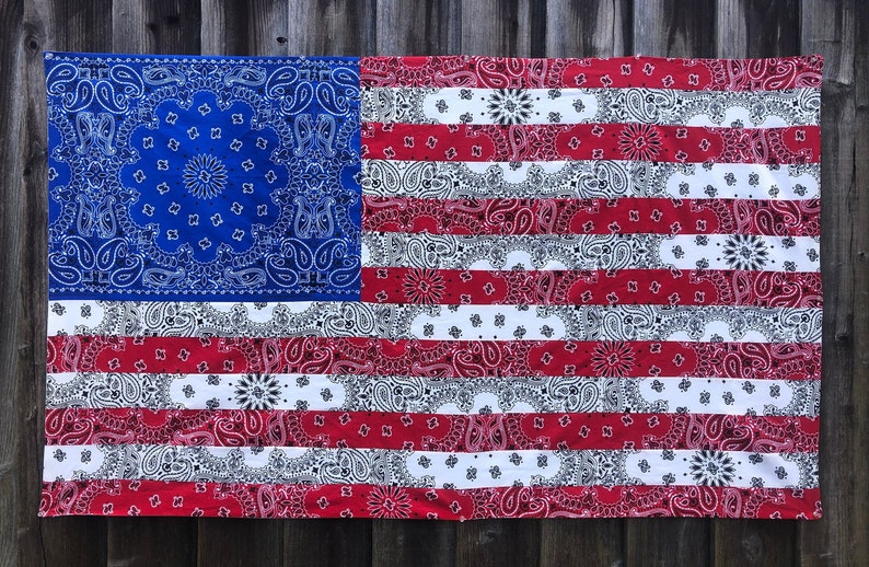May include: A large American flag made from red, white, and blue bandanas. The flag is hanging on a wooden fence.