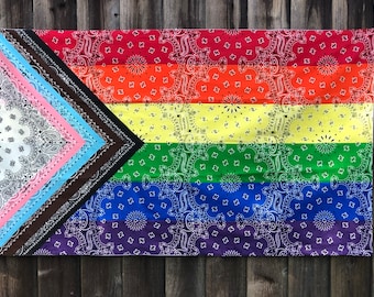 May include: A rainbow pride flag made of bandanas. The flag is made of six horizontal stripes of red, orange, yellow, green, blue, and purple. The stripes are all made of different patterned bandanas. The flag is hanging on a wooden fence.