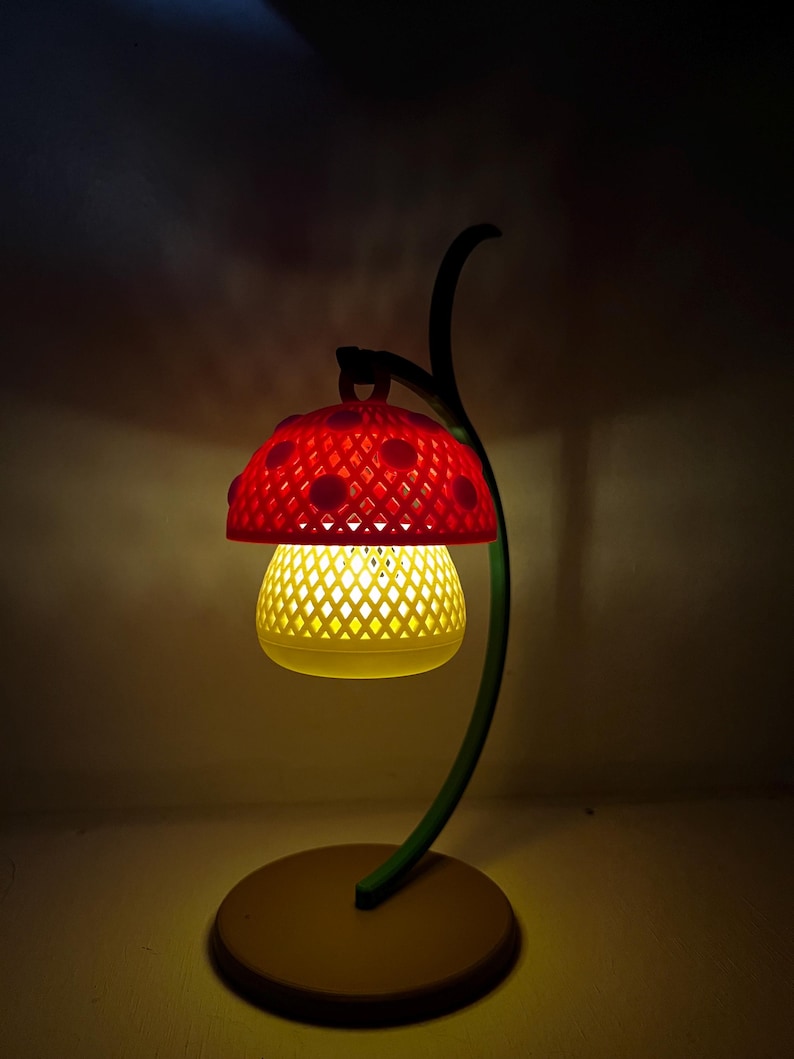 May include: A whimsical mushroom-shaped lamp with a red, perforated cap and a yellow, diamond-patterned shade. The lamp has a green, curved stem and a round, tan base. The lamp is lit, casting a warm glow.