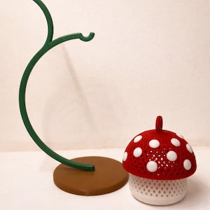 May include: A whimsical mushroom-shaped decorative item with a red cap dotted with white spots. The mushroom sits next to a green, curved stand with a brown base. The mushroom has a white, lattice-like base.