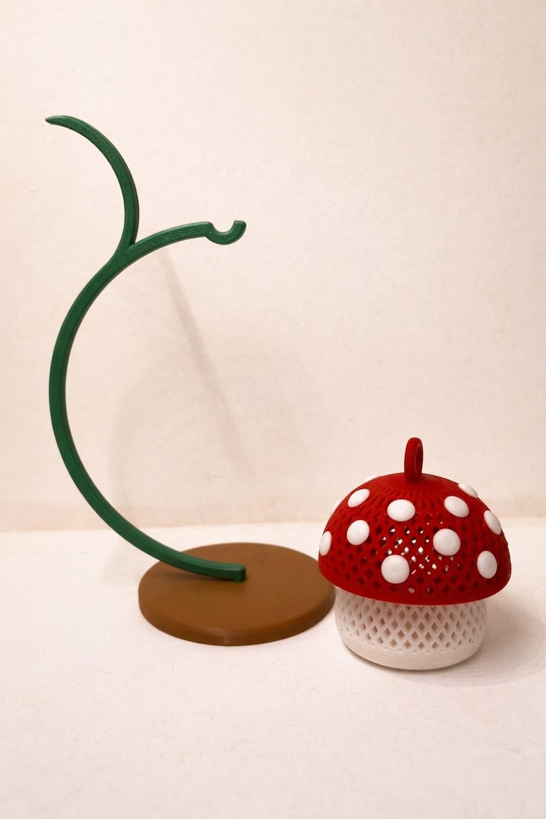 May include: A whimsical mushroom-shaped decorative item with a red cap dotted with white spots. The mushroom sits next to a green, curved stand with a brown base. The mushroom has a white, lattice-like base.