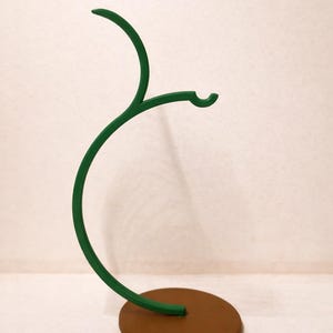 May include: A decorative, green metal stand with a brown circular base. The stand has a curved, branch-like design with a hook at the end. The stand is designed to hold small items.