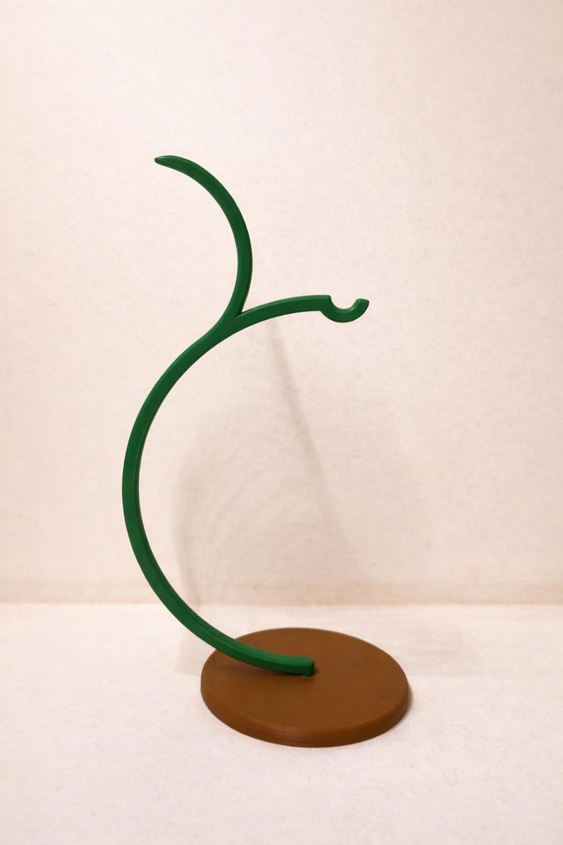 May include: A decorative, green metal stand with a brown circular base. The stand has a curved, branch-like design with a hook at the end. The stand is designed to hold small items.