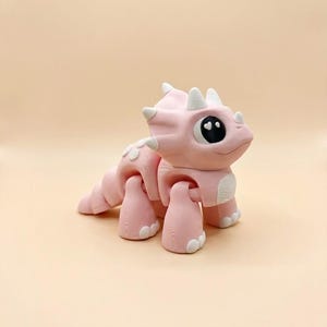 May include: A pink triceratops figurine with white accents on its horns, frill, and feet. The dinosaur has large, expressive eyes with heart-shaped pupils. The toy is made of a smooth material and has a friendly, cartoonish appearance.