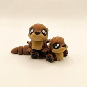 May include: Two brown otter figurines with tan bellies and large, expressive eyes. One otter is sitting upright, while the other is lying down. The otters have detailed paw pads and a segmented tail. The figurines are likely 3D printed.
