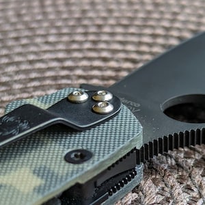 May include: Close-up of a black pocket knife with a green and brown camouflage handle. The knife has a partially open blade and three silver screws on the handle.