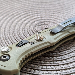 May include: A green folding knife with a black handle and a silver clip. The knife has a small silver screw on the side.