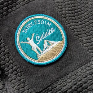 Puede incluir: Primer plano de una chaqueta polar gris oscuro con un parche bordado circular. El parche presenta el texto "TATRY, 2301M Świnica" en turquesa y blanco, con un gráfico de esquiador y una ilustración de montaña en dorado.