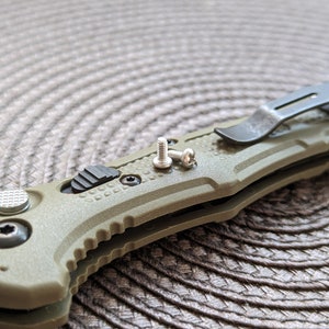 Puede incluir: Un cuchillo plegable verde con un clip de bolsillo negro y dos tornillos plateados. El cuchillo está sobre una superficie tejida marrón.