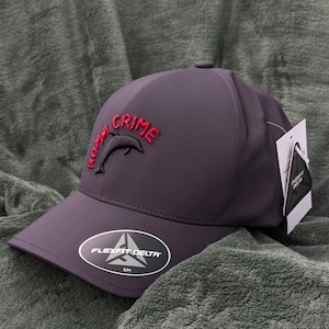 May include: A dark grey baseball cap with a red embroidered dolphin and the text "Miami Crime". The cap has a black Flexfit Delta logo on the underside of the brim.