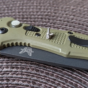 以下が含まれることがあります： Benchmade のロゴが付いた緑と黒の折りたたみナイフのクローズアップ。ナイフに2本の小さな銀色のネジがある。