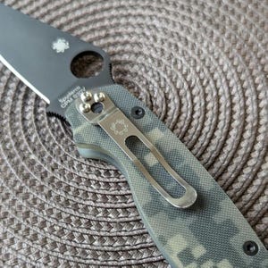 May include: A Spyderco CPM S90V knife with a black blade and a camouflage handle. The knife has a pocket clip and a Spyderco logo on the blade.