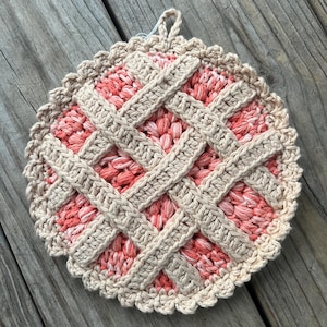 May include: A crocheted pot holder shaped like a pie, with a beige and pink colour scheme. The pot holder has a scalloped edge.