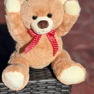 May include: A light brown teddy bear with a red and white polka dot bow tie. The bear is sitting on a black wicker surface with its arms raised.