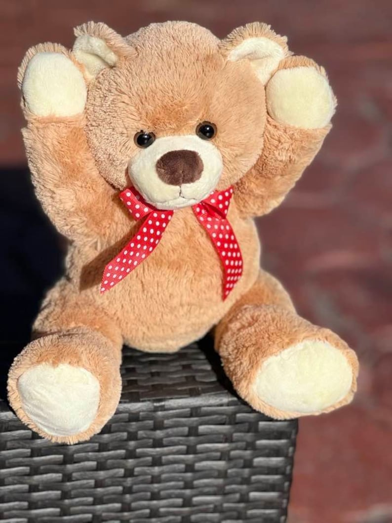 May include: A light brown teddy bear with a red and white polka dot bow tie. The bear is sitting on a black wicker surface with its arms raised.