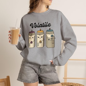 May include: A heather grey crewneck sweatshirt featuring the word Volatile in black script above a graphic of three cartoon-style laboratory instruments with faces. The centre design includes three distinct scientific containers in shades of brown.