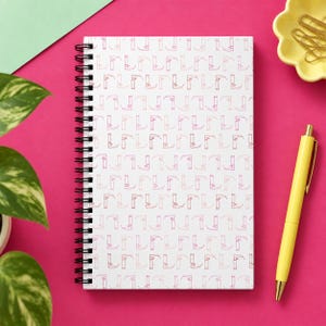 May include: A white spiral-bound notebook featuring a repeating pattern of small pink scythe icons. The notebook has a black metal coil binding along the left side and a clean, minimalist white cover with the pink graphic print throughout.
