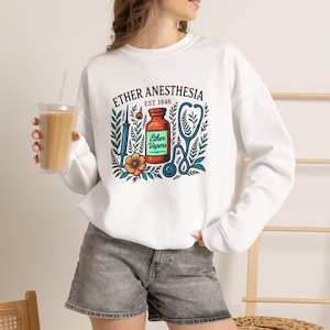 May include: A white long-sleeve sweatshirt featuring a graphic print with the text ETHER ANESTHESIA EST. 1846. The design includes a central orange bottle labelled Ether Vapors, a blue syringe, a blue stethoscope, and colourful floral foliage.