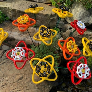 May include: A bouquet of colorful metal flowers made from repurposed water valve handles. The flowers are yellow, red, and orange with black accents. The flowers are arranged in a vase and are sitting on a bed of rocks.