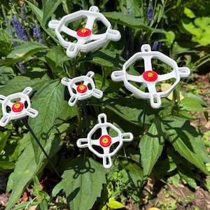 May include: White and red decorative garden stakes with a unique design. The stakes have a white frame with a red center and are placed among green foliage and flowers. These stakes add a decorative touch to any garden.
