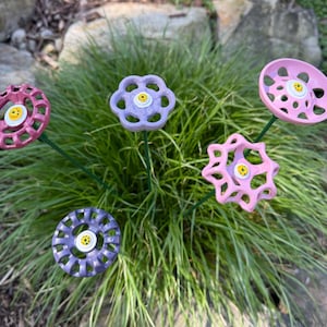 May include: Five colorful metal flower decorations made from repurposed plumbing parts. The flowers are pink, purple, and lavender with yellow centers. The flowers are arranged in a grassy area.
