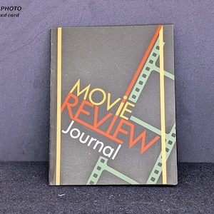 May include: A movie review journal with a dark grey cover. The title "MOVIE REVIEW Journal" is displayed in yellow, orange, and white. The cover also features a design of green and orange film strips. A yellow vertical stripe is on the left side.