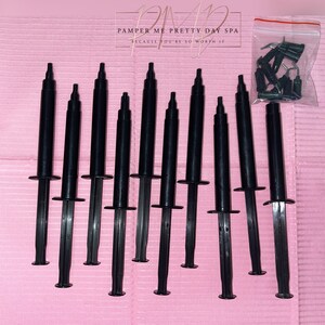 May include: Ten black plastic syringes with a clear tip, arranged in a row on a pink surface. The text "Pamper Me Pretty Day Spa" is visible in the background.