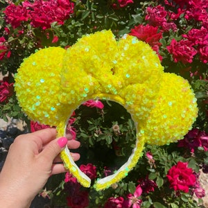 May include: A yellow sequined headband with large, rounded ears. The headband is covered in bright yellow sequins, with a white interior. The ears are also covered in sequins. The background features red roses.