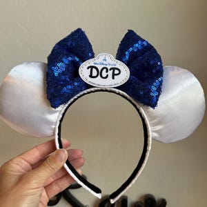 May include: White satin mouse ears with a navy blue sequined bow and a black headband. The bow features a white oval with the text "DCP" and "Walt Disney World". The ears are held by a hand.