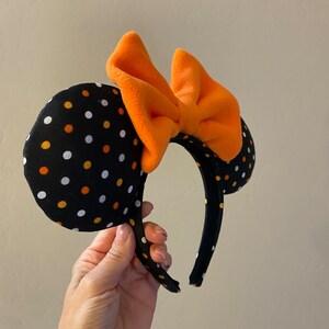 May include: Black and white polka dot Minnie Mouse ears headband with an orange bow. The headband is made of fabric and has a black elastic band.