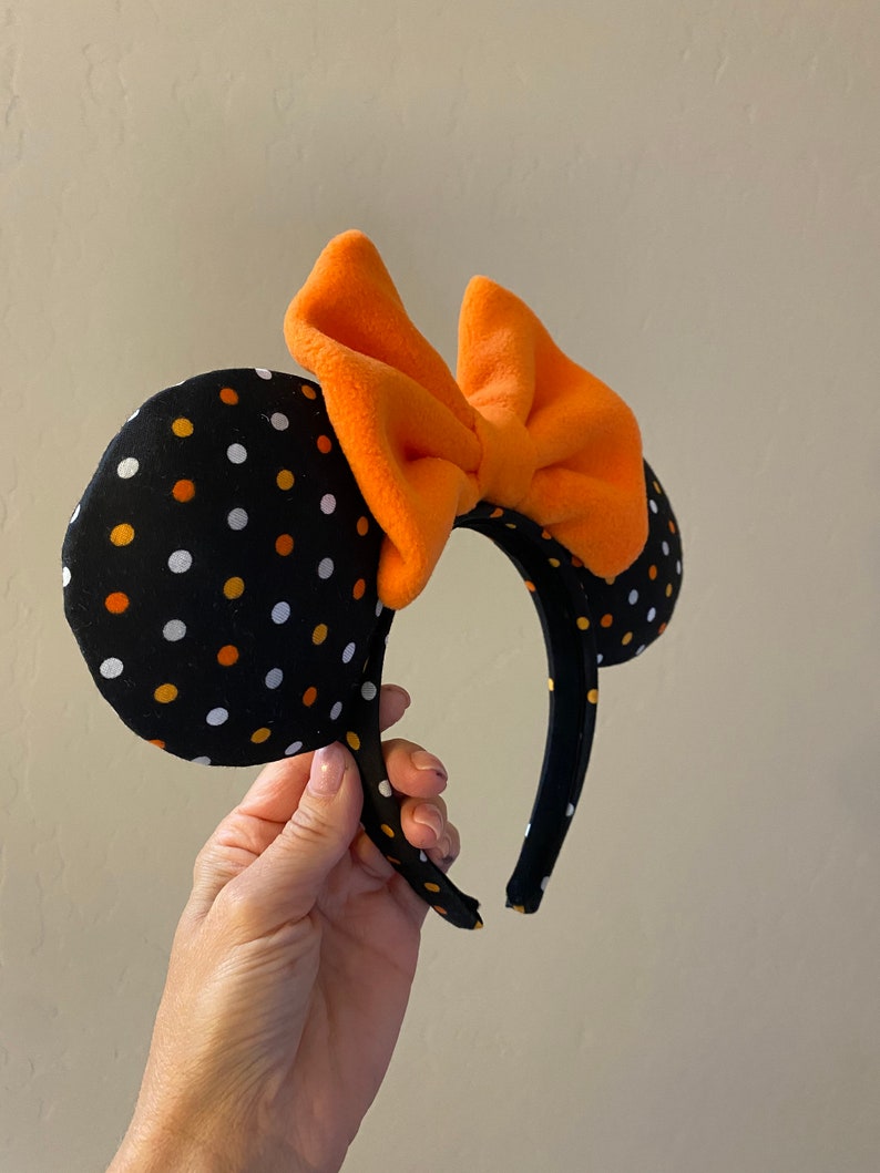 May include: Black and white polka dot Minnie Mouse ears headband with an orange bow. The headband is made of fabric and has a black elastic band.