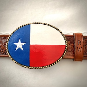 May include: Oval belt buckle featuring the Texas state flag design. The buckle has a blue field with a white star, a white stripe, and a red stripe. The buckle is attached to a brown leather belt with carved details.