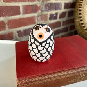 May include: A hand-painted ceramic owl figurine. The owl has a white body with black and orange details. The owl is egg-shaped and has a stylized design. The figurine is sitting on a red book.