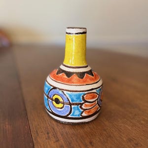 May include: A small, hand-painted ceramic vase with a rounded body and a slender neck. The vase features a vibrant color palette, including yellow, orange, blue, and black. The design includes geometric and floral patterns, creating a unique decorative piece.