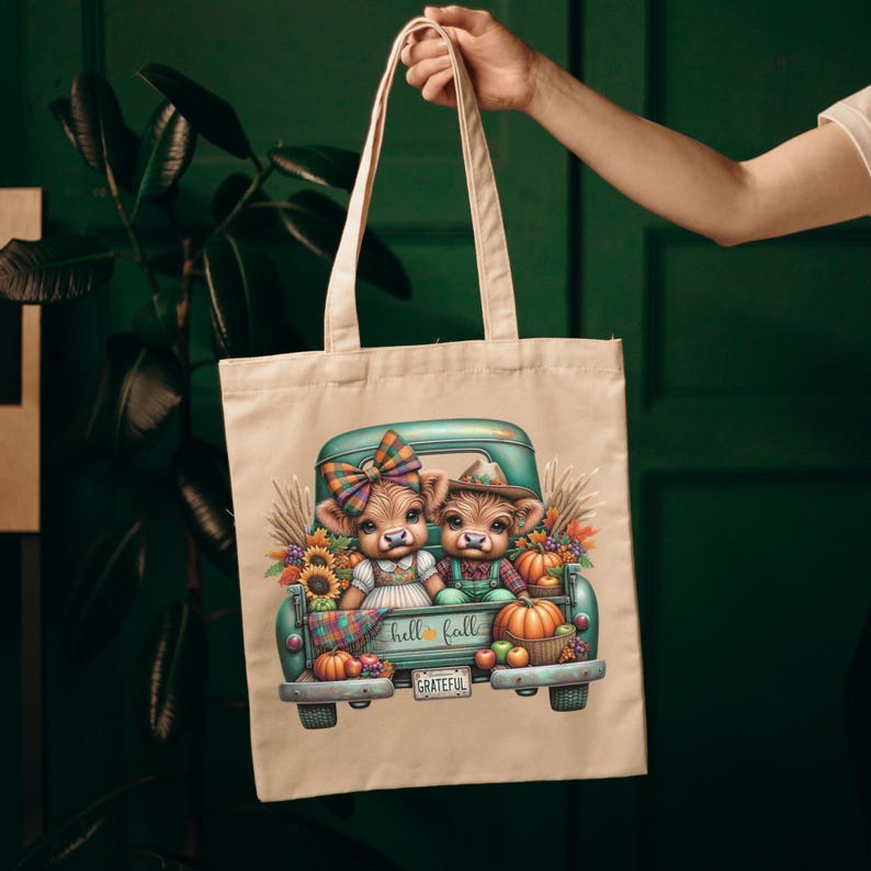 May include: A beige tote bag featuring a teal pickup truck filled with pumpkins, sunflowers, and two cartoon cows. The truck bed has the words "hello fall" and "grateful". The cows are wearing fall-themed outfits.