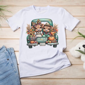 May include: White t-shirt featuring a teal truck bed filled with pumpkins, sunflowers, and two cartoon cows. The cows are dressed in fall attire. The text "hello fall" and "grateful" are visible on the design.
