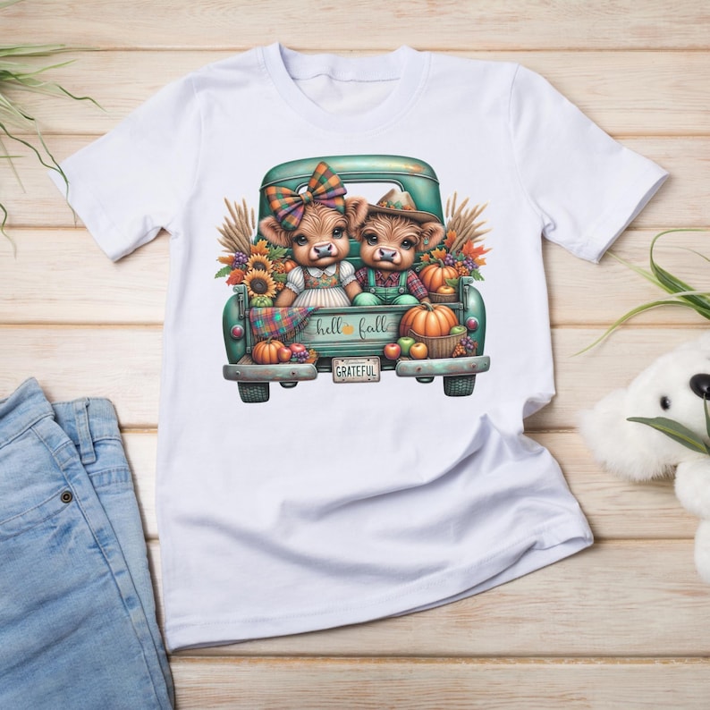 May include: White t-shirt featuring a teal truck bed filled with pumpkins, sunflowers, and two cartoon cows. The cows are dressed in fall attire. The text "hello fall" and "grateful" are visible on the design.