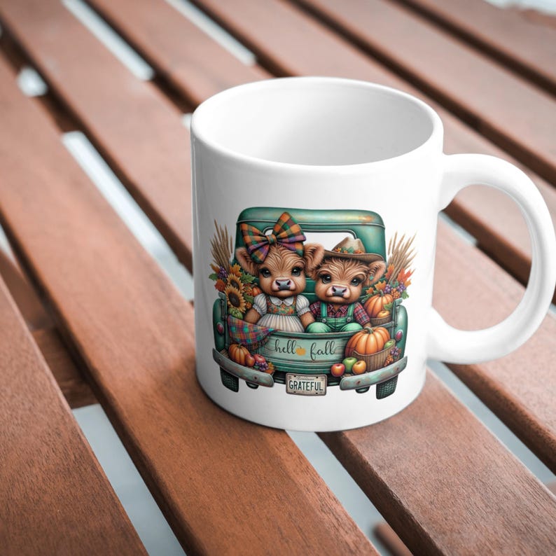May include: White ceramic mug featuring a teal pickup truck bed with two cartoon cows. The truck bed is filled with pumpkins, sunflowers, and fall foliage. The text "hello fall" is on the truck bed, and "grateful" is on the bumper.
